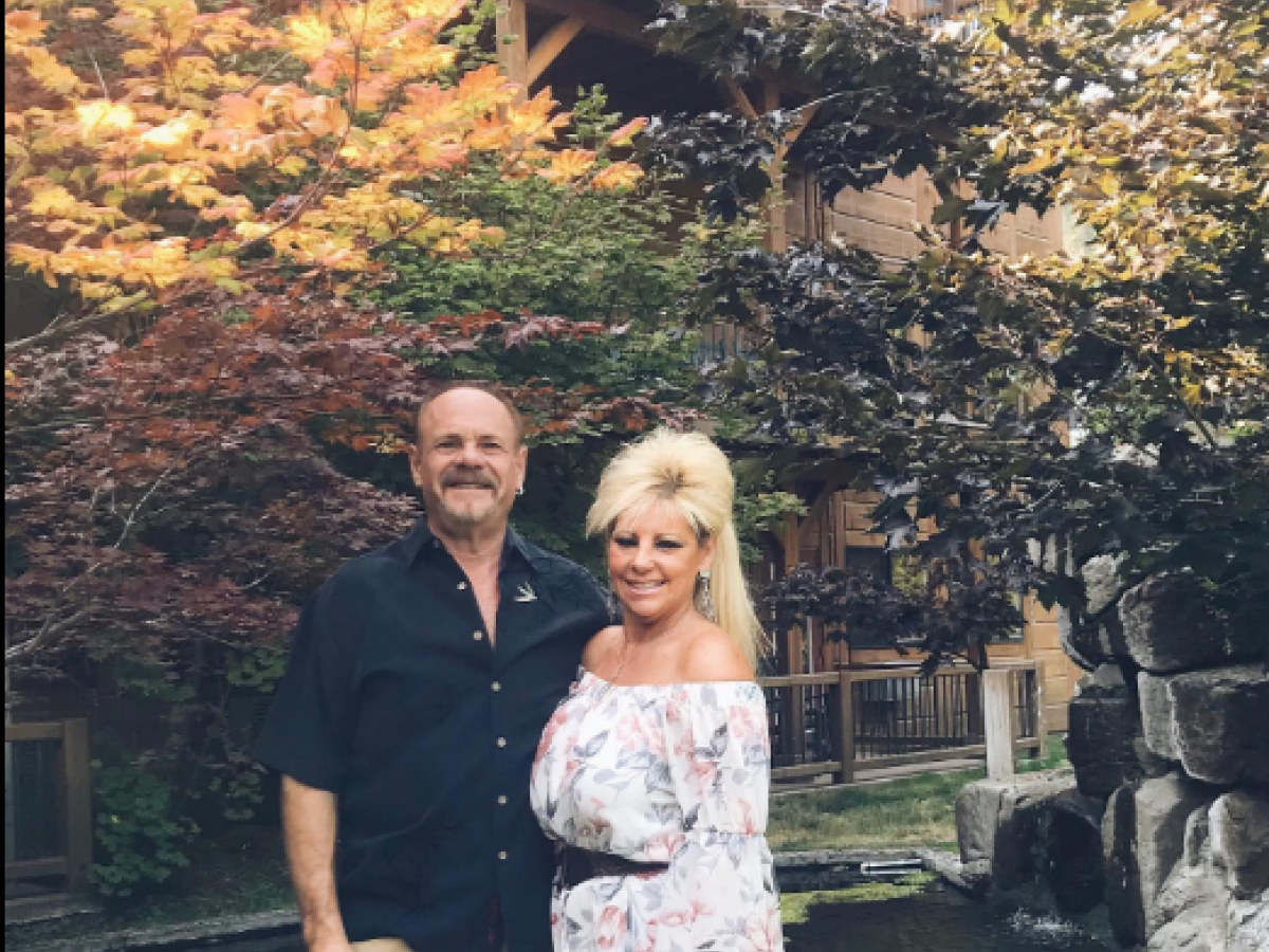 A man and a woman stand together in a garden with colorful trees and a small pond, in front of a wooden building.