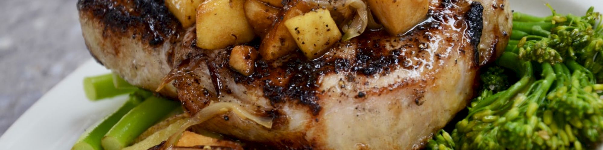 Grilled pork chop topped with diced apples, served with roasted garlic, broccolini, and roasted potato on the side, garnished with herbs.