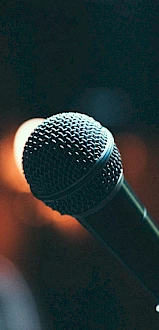 The image shows a close-up of a microphone against a blurred, dimly lit background, suggesting a performance or speaking event.
