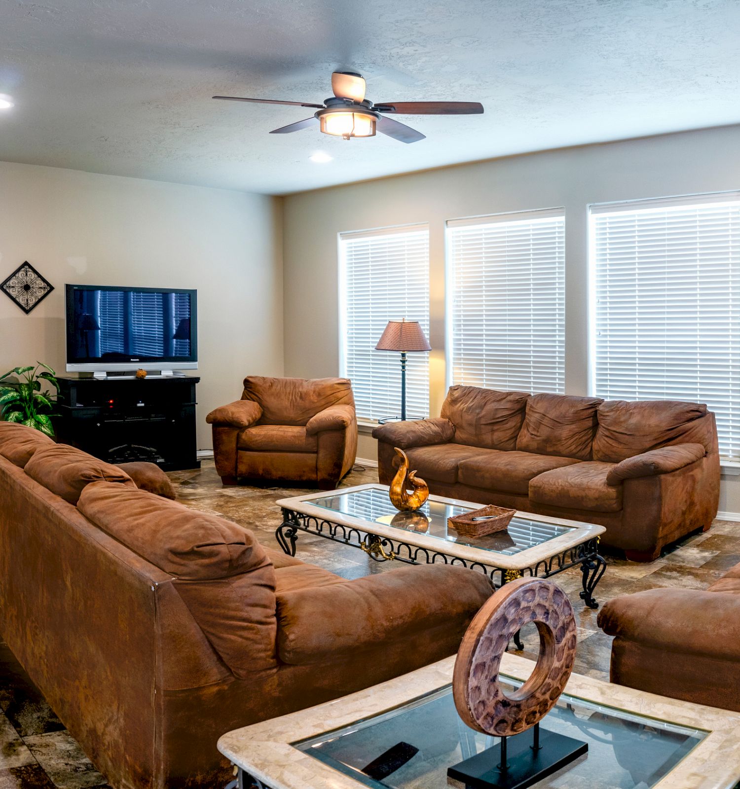 A cozy living room with brown sofas, a ceiling fan, a wall-mounted TV, and decorative items. The room has large windows with blinds and a "HOME" sign.