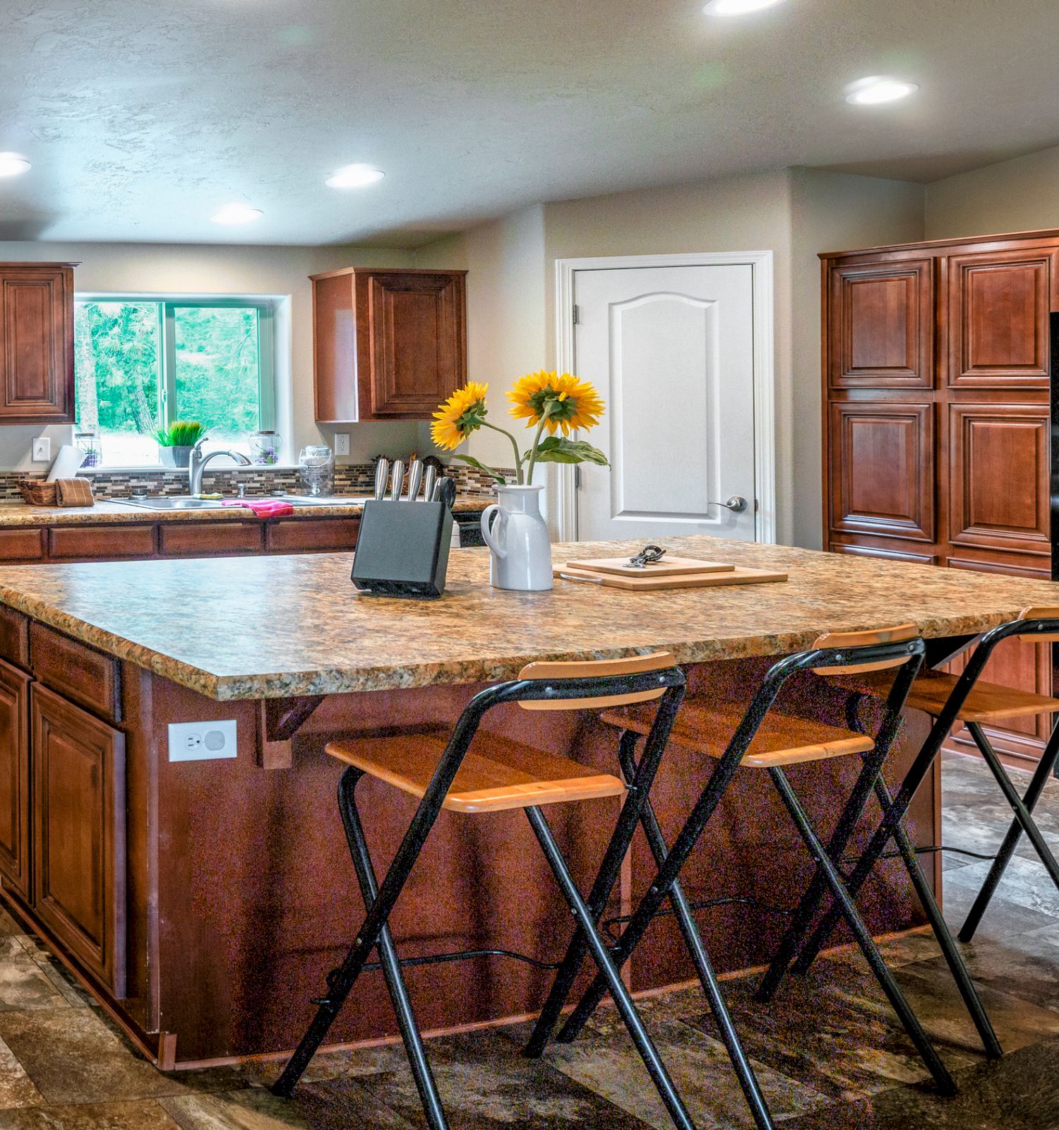 A spacious kitchen with wooden cabinets, an island with three stools, black appliances, and a vase of sunflowers on the countertop ends the sentence.