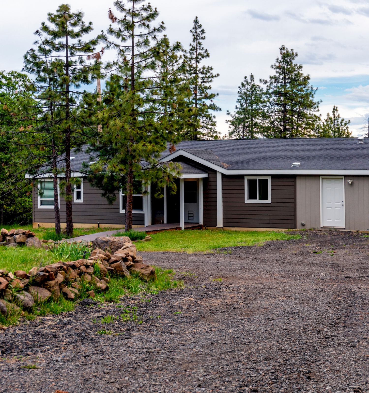 A single-story house in a rural setting surrounded by trees, with a gravel driveway leading up to the entrance and a stone-lined pathway.