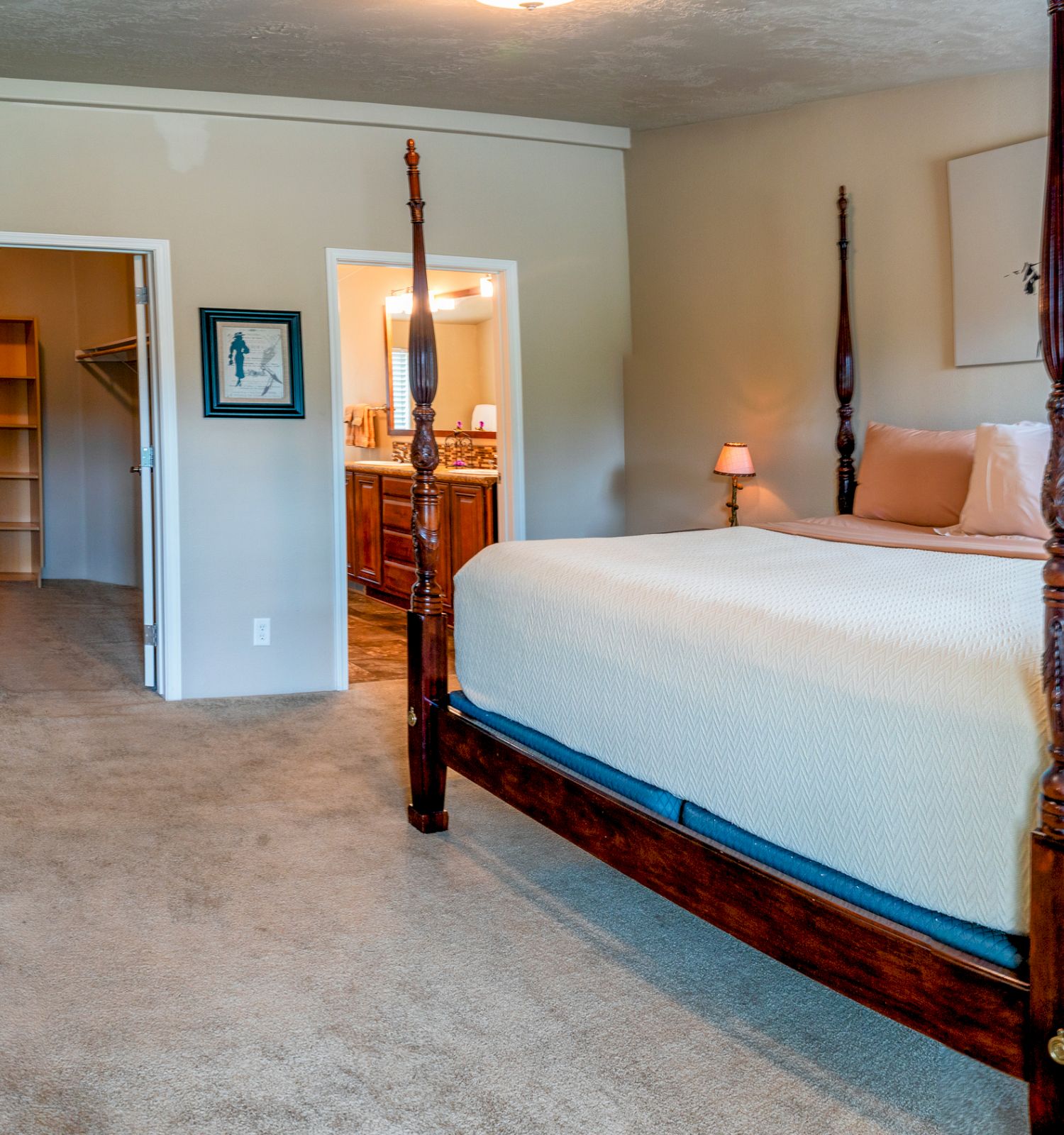 This image shows a spacious bedroom with a four-poster bed, nightstands with lamps, a dresser, a pink chair, carpeted floor, and an open walk-in closet.