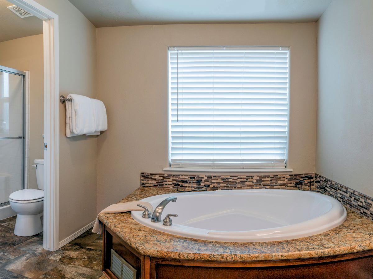 The image shows a bathroom with a round bathtub, a window with blinds, a towel on a rack, and a toilet and shower area visible through an open door.