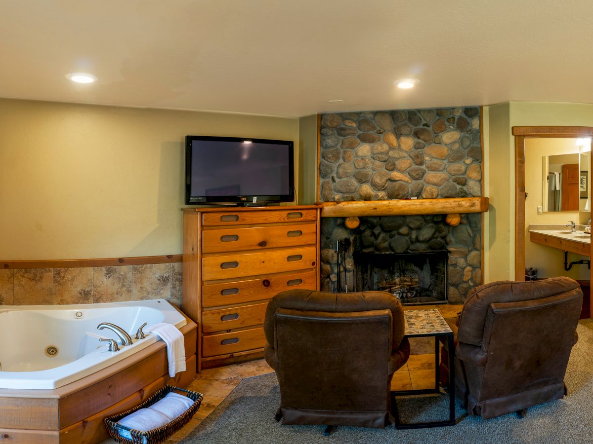 A cozy room with a fireplace, TV, dresser, two armchairs, and a Jacuzzi tub, next to a small table. There’s a hallway leading to a bathroom.