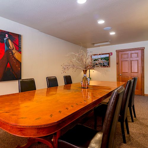 A conference room with a wooden table, eight black chairs, a decorative centerpiece, a painting on the wall, a lamp, and a closed door.