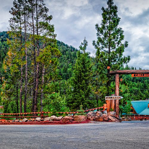 The image shows a lodge entrance surrounded by trees and mountains, featuring a wooden archway with signage and a small road in the foreground.