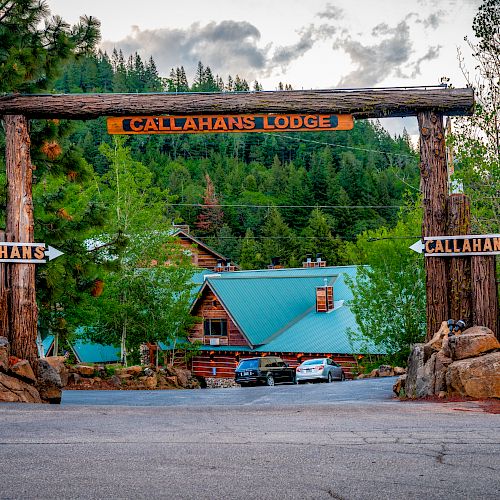 A wooden archway with the sign "Callahan's Lodge" stands at the entrance to a lodge amidst lush greenery and pine trees.