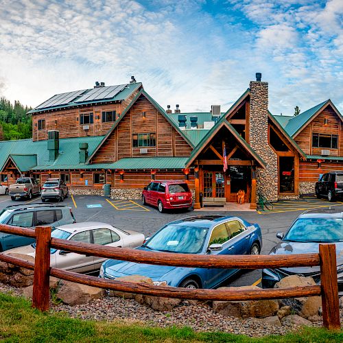 The image shows a large wooden lodge with a green roof, cars parked in front, surrounded by lush greenery and a wooden fence, under a partly cloudy sky.