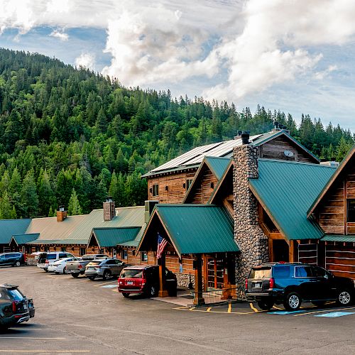 This image shows a rustic lodge with green roofs and wooden facades set against a forested hill. Parked cars are visible in front of the buildings.