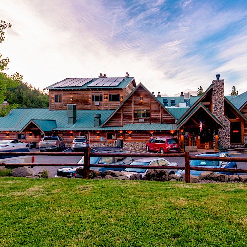 The image depicts a rustic lodge with green roofs, set against a backdrop of trees, with cars parked in front and a grassy area in the foreground.