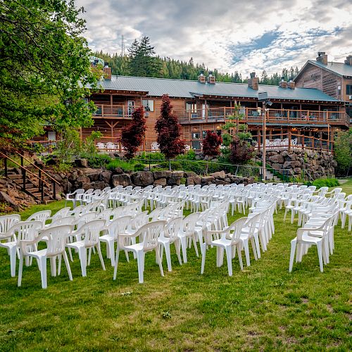 An outdoor venue with rows of white chairs facing a small wooden stage, set against a backdrop of rustic buildings and trees under a partly cloudy sky.