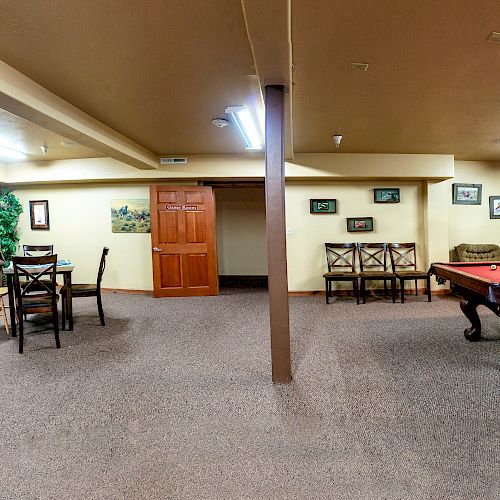 The image shows a spacious, well-lit recreation room with a pool table, cozy sofas, chairs, tables, wall art, and a potted plant.
