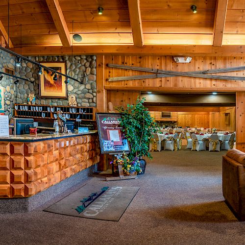 The image shows the interior of a rustic lodge with a wooden reception desk, seating area, and a dining hall set up with tables and chairs.
