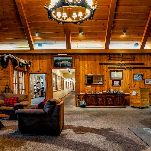 A cozy lodge with wooden interiors features leather sofas, a chandelier, taxidermy decor, and multiple framed pictures hanging on the walls.