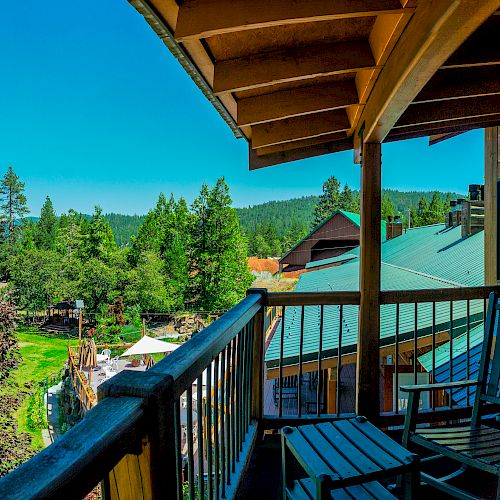 A balcony with two wooden chairs overlooks a scenic landscape of trees, rooftops, and a clear blue sky in a peaceful, forested area.