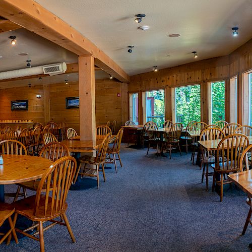 This image shows a cozy dining area with wooden tables and chairs, surrounded by large windows letting in natural light.