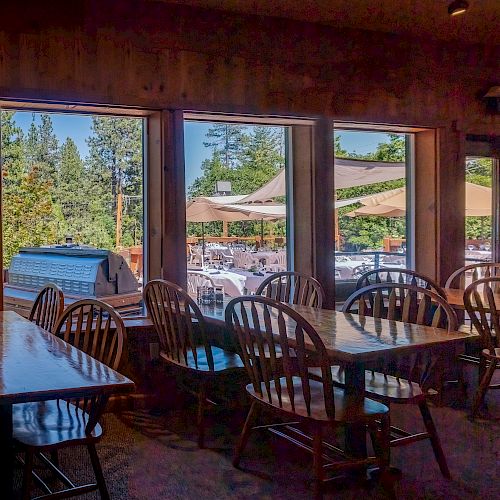 This image shows an indoor dining area with wooden tables and chairs, and large windows overlooking an outdoor patio with umbrellas and seating in a forested area.