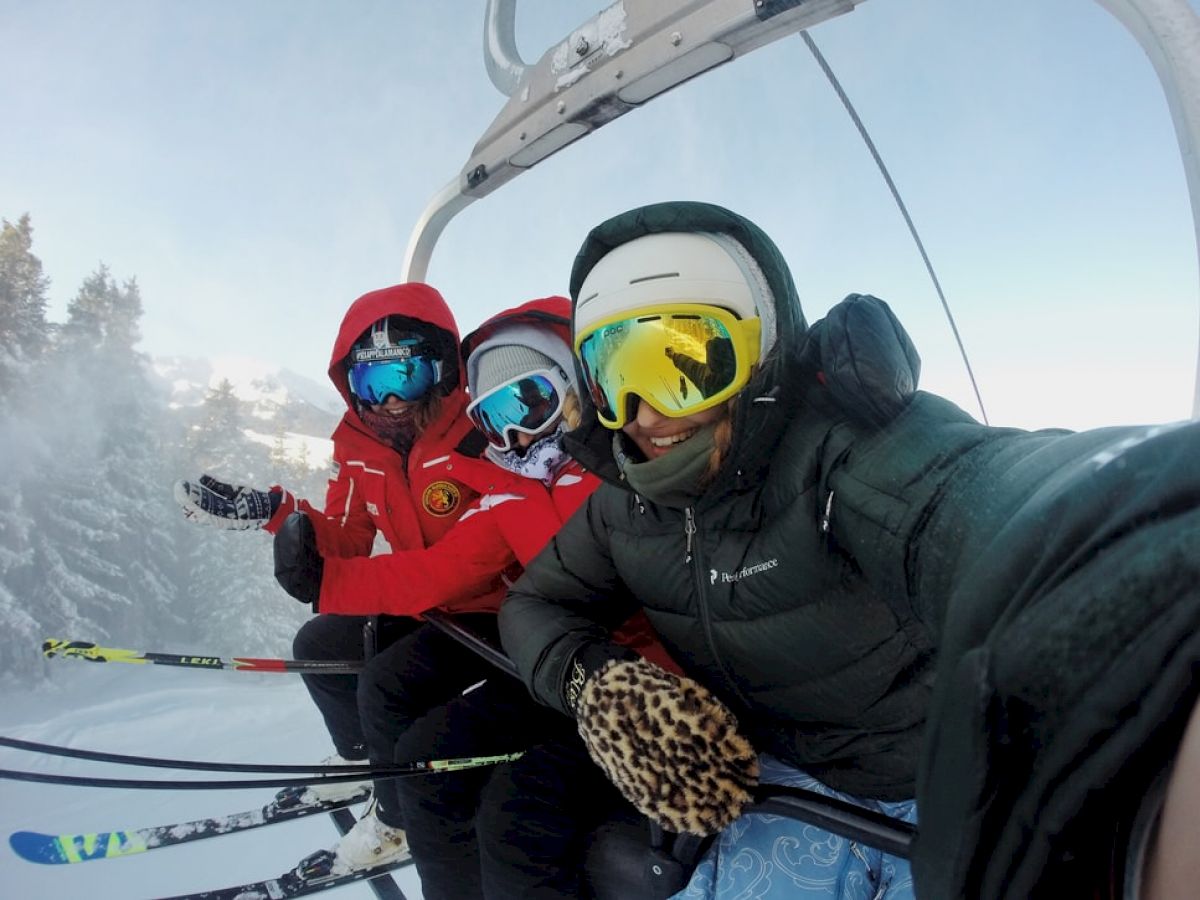 Three people in winter gear and ski goggles are on a ski lift, holding ski poles with snowy trees in the background, posing for a selfie.