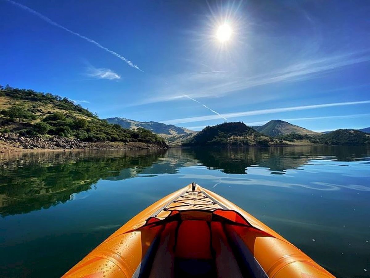 A kayak on a calm lake with mountains in the background and a bright sun in the blue sky, creating a peaceful and scenic outdoor view.