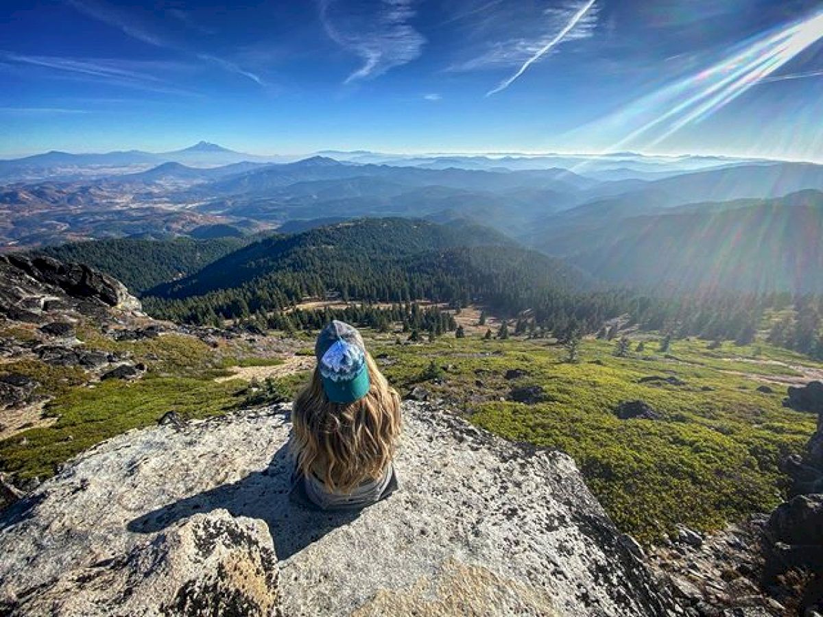 Person sits on a rocky ledge, overlooking a vast mountainous landscape with clear blue skies and lush greenery, under a bright sun.