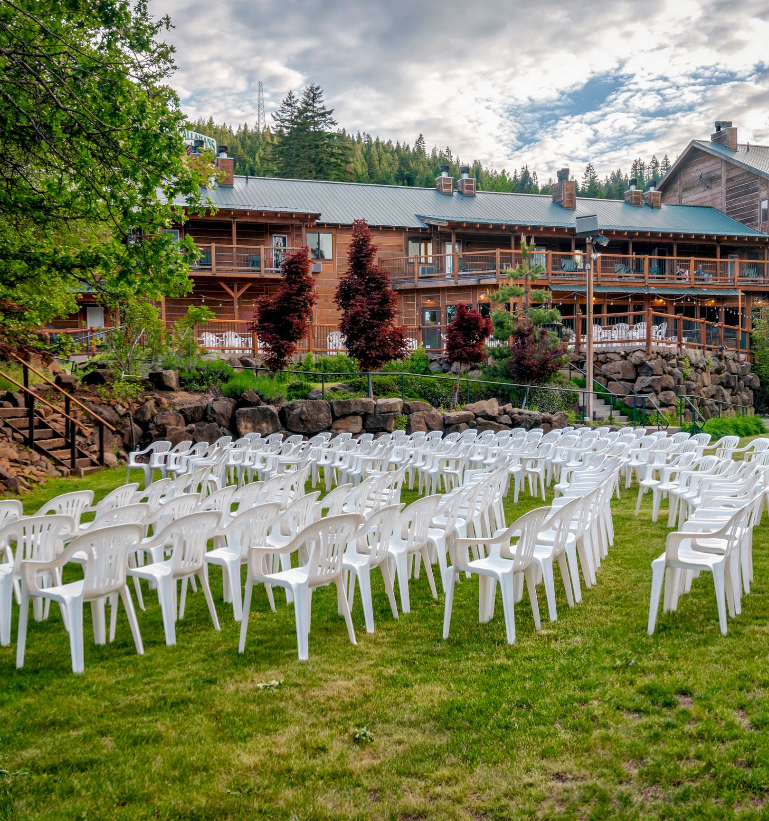 Rows of white chairs set up on a lawn, facing a small wooden gazebo and a large building in the background with greenery and trees surrounding.