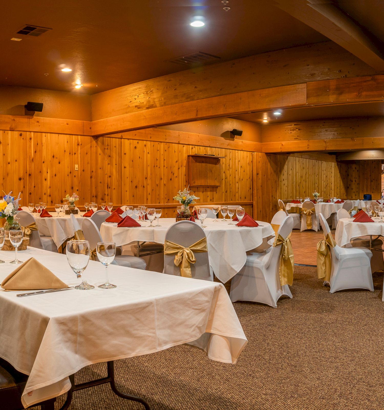 An elegantly decorated banquet hall with white tablecloths, red napkins, and gold chair sashes, ready for an event.