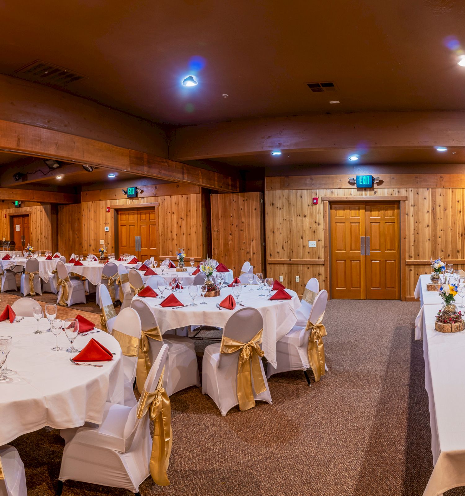 A banquet hall is set up for an event with round tables having white tablecloths, red napkins, and gold chair sashes, all arranged neatly in the room.
