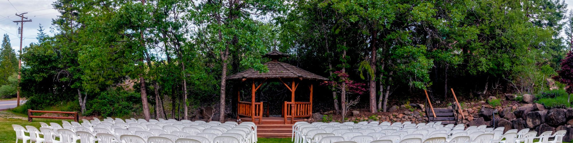An outdoor wedding setup with rows of white chairs facing a wooden gazebo in a garden surrounded by trees and greenery.
