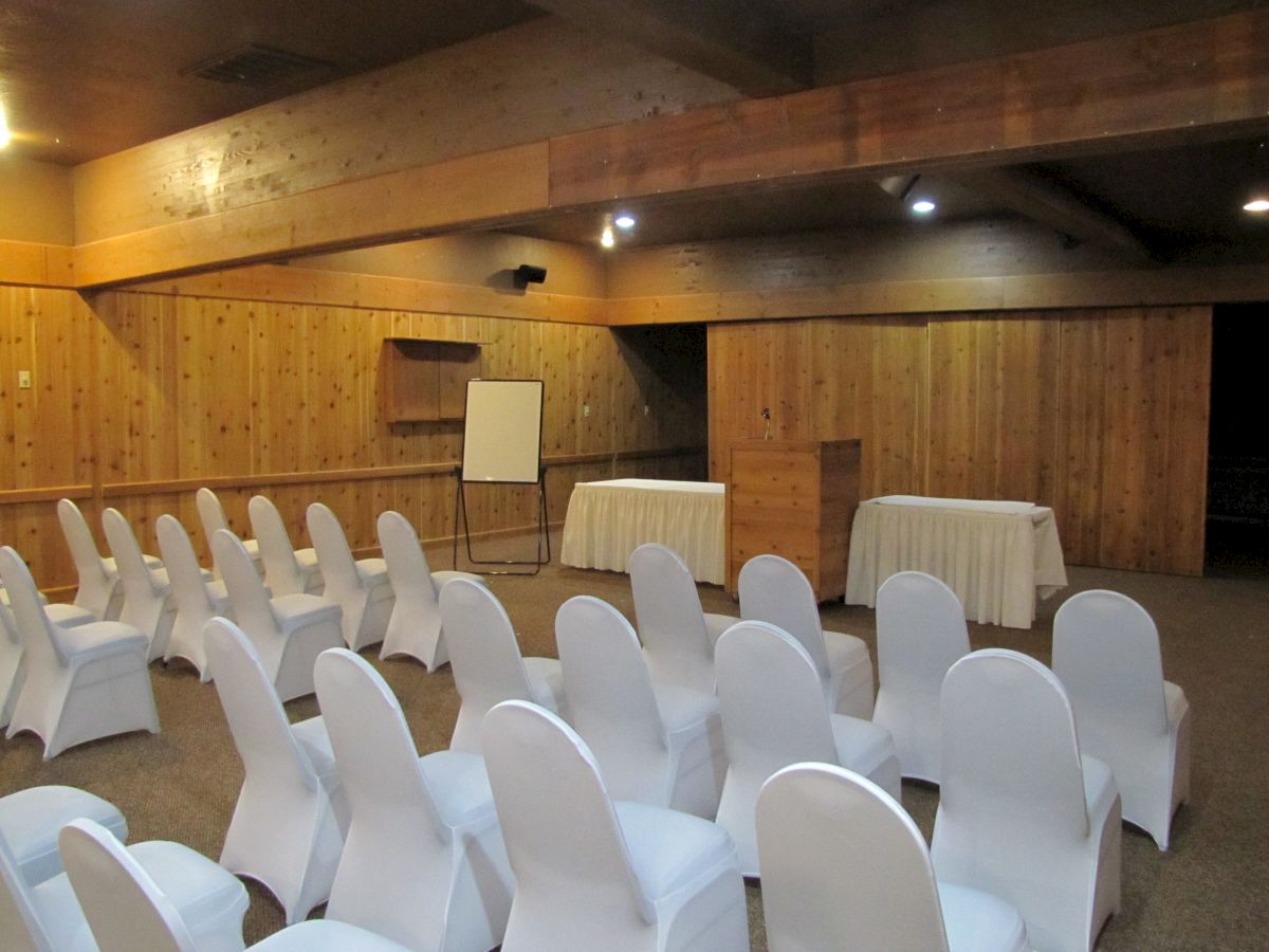 This image shows a conference room setup with rows of white chair covers, a whiteboard, and two tables covered with white tablecloths, in a wooden interior.
