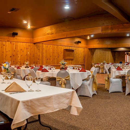 The image shows a banquet hall with tables set for an event, featuring white tablecloths, folded napkins, and chair covers with gold sashes.