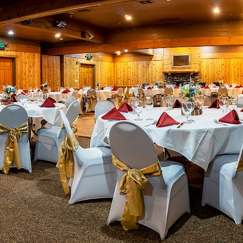 The image shows a banquet hall with round tables covered in white tablecloths, red napkins, and chairs adorned with gold sashes.