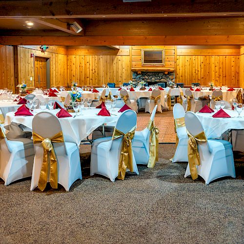 The image shows a decorated banquet hall with round tables set for an event, featuring white chair covers with gold bows and red napkins.