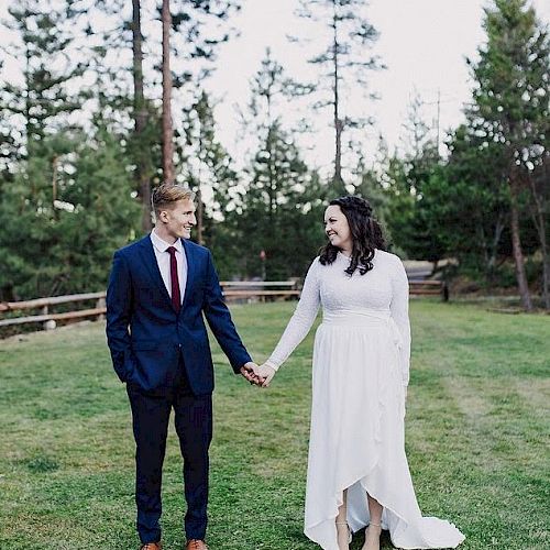 A couple, dressed in formal attire, stands hand in hand on a grassy area outdoors with trees in the background.