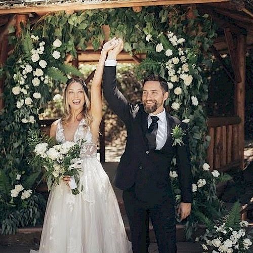 A couple celebrates in front of a flower-adorned gazebo, with the bride holding a bouquet and both raising their hands in happiness.
