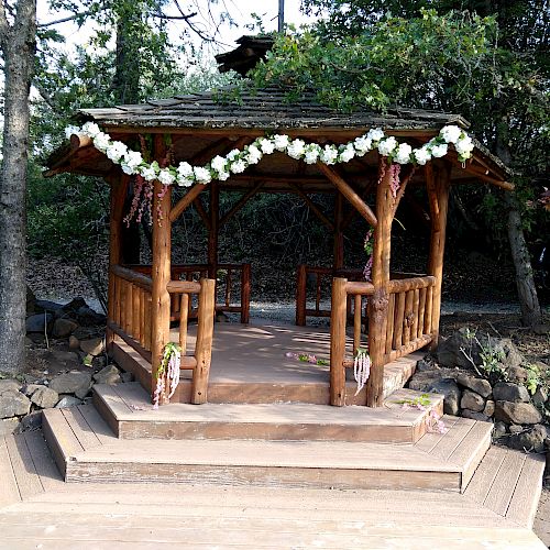 A wooden gazebo is adorned with a garland of white flowers, set in a wooded area with leafy surroundings and stone steps leading up to it.