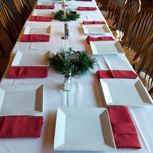 A long, rectangular dining table is set up with white plates, red napkins, and greenery decorations, ready for a meal or event.