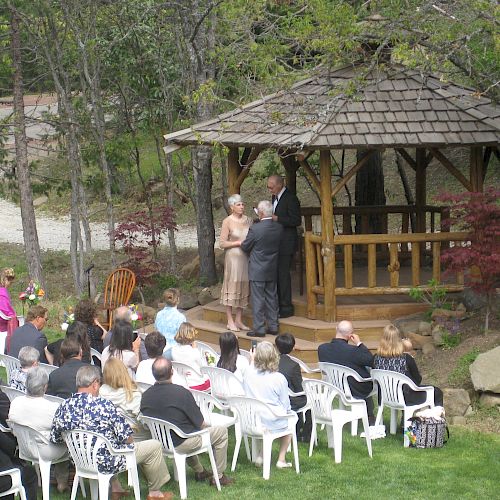 A wedding ceremony taking place outside with attendees seated in white chairs, facing a couple and officiant under a gazebo.