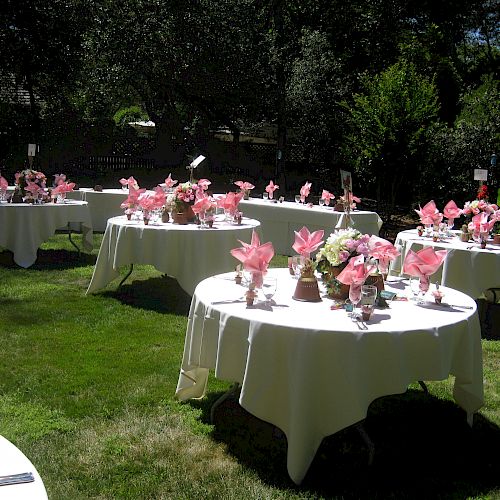 Tables with white tablecloths and pink napkins in an outdoor garden setting, decorated for an event with flowers as centerpieces.