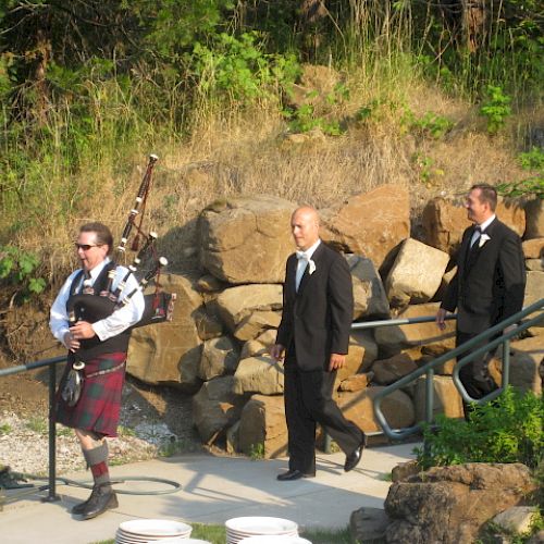 A man in a kilt plays bagpipes, leading two men in suits down a path bordered by rocks and greenery.