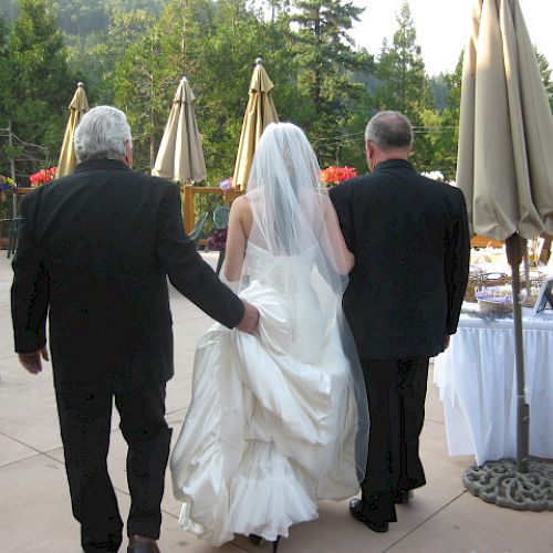 A bride in a white wedding dress with a veil walks between two men in black suits, outdoors with a scenic background, ending the sentence.