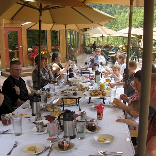 People are gathered around a long outdoor dining table with food, drinks, and umbrellas, enjoying a meal together.