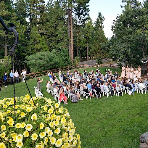 An outdoor wedding ceremony with guests seated on a lawn, surrounded by trees and flowers, with the bridal party and officiant at the front.