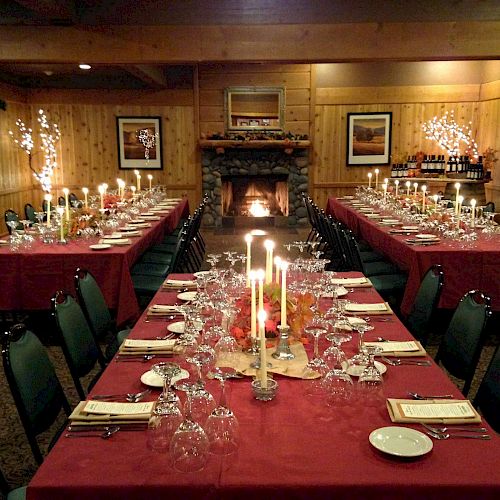 The image shows a festive dining room with red tablecloths, elaborate table settings, candles, and a lit fireplace in a cozy wooden interior.