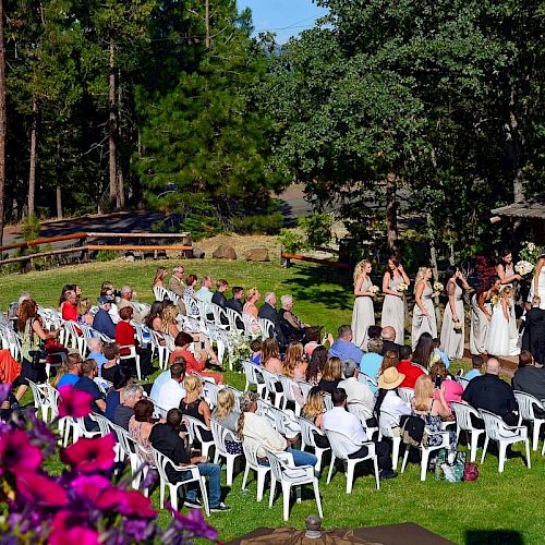 An outdoor wedding ceremony is taking place with guests seated on white chairs, surrounded by lush greenery and trees, under a clear blue sky.