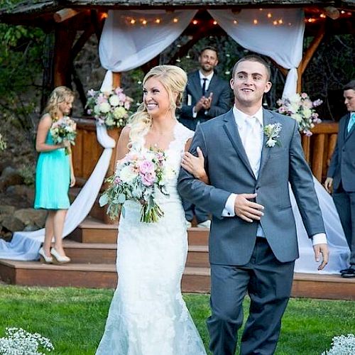 A bride and groom are walking down the aisle outdoors, smiling. Bridesmaids and groomsmen stand near a decorated gazebo in the background.