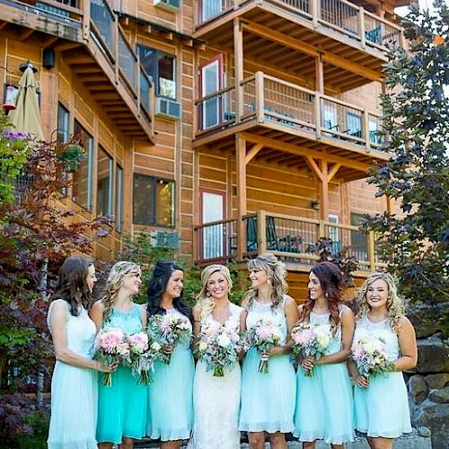 A bride stands with six bridesmaids, each holding bouquets, in front of a rustic wooden building with multiple balconies.