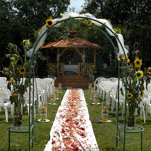 The image depicts an outdoor wedding setup with a flower-lined aisle, white chairs, and a pergola decorated with sunflowers in a garden setting.