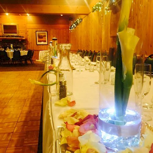 The image shows a decorated banquet hall with a long dining table, featuring a vase with flowers and scattered petals, set against a wooden interior.