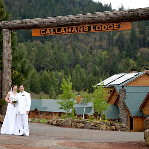 A couple dressed in wedding attire stands in front of Callahan's Lodge, nestled in a scenic wooded area with log buildings in the background.